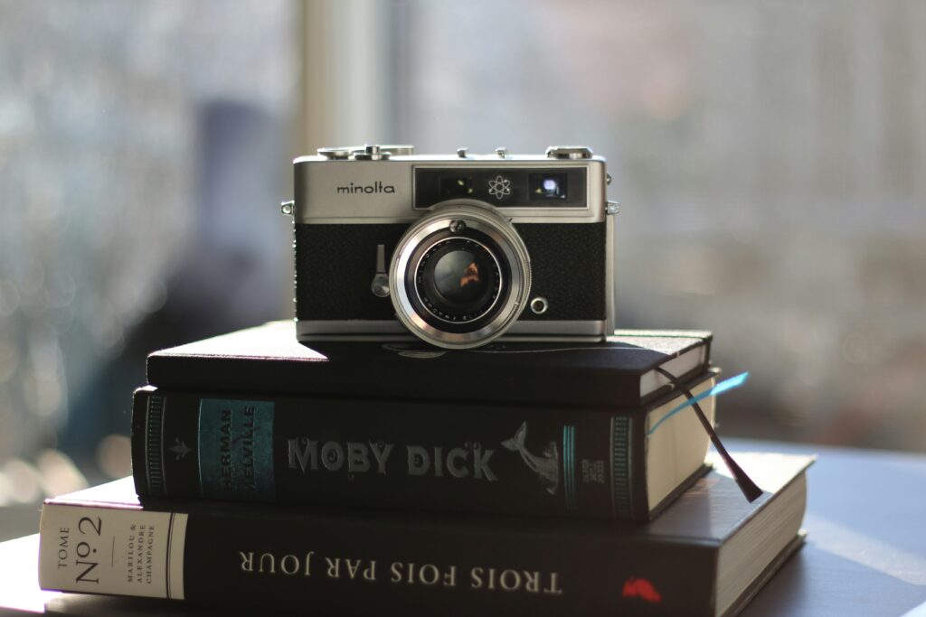 A vintage Minolta camera is centred on a stack of hardcover books, including "Moby Dick."