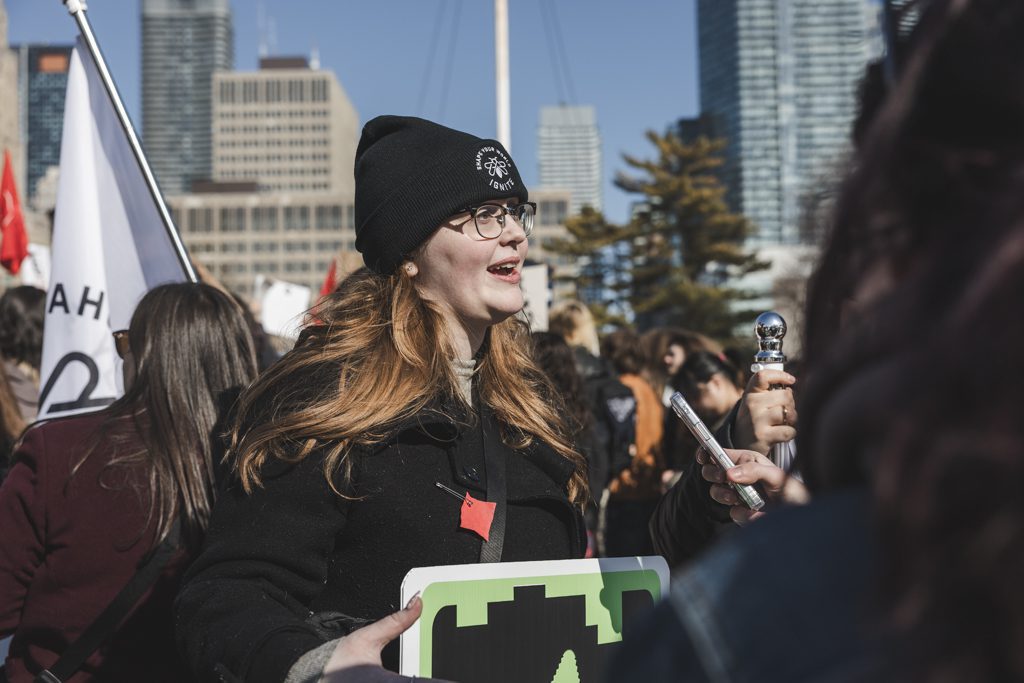A woman wearing a black coat and a beanie speaks to others at a rally. She is holding a sign and surrounded by people and flags.