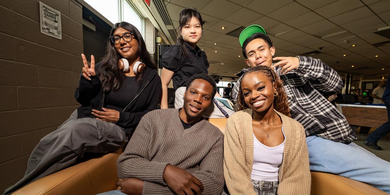Five young adults sit and pose together indoors, smiling and making peace signs. Some wear casual clothing and headphones. The background shows a lounge area with tables, lights, and windows.