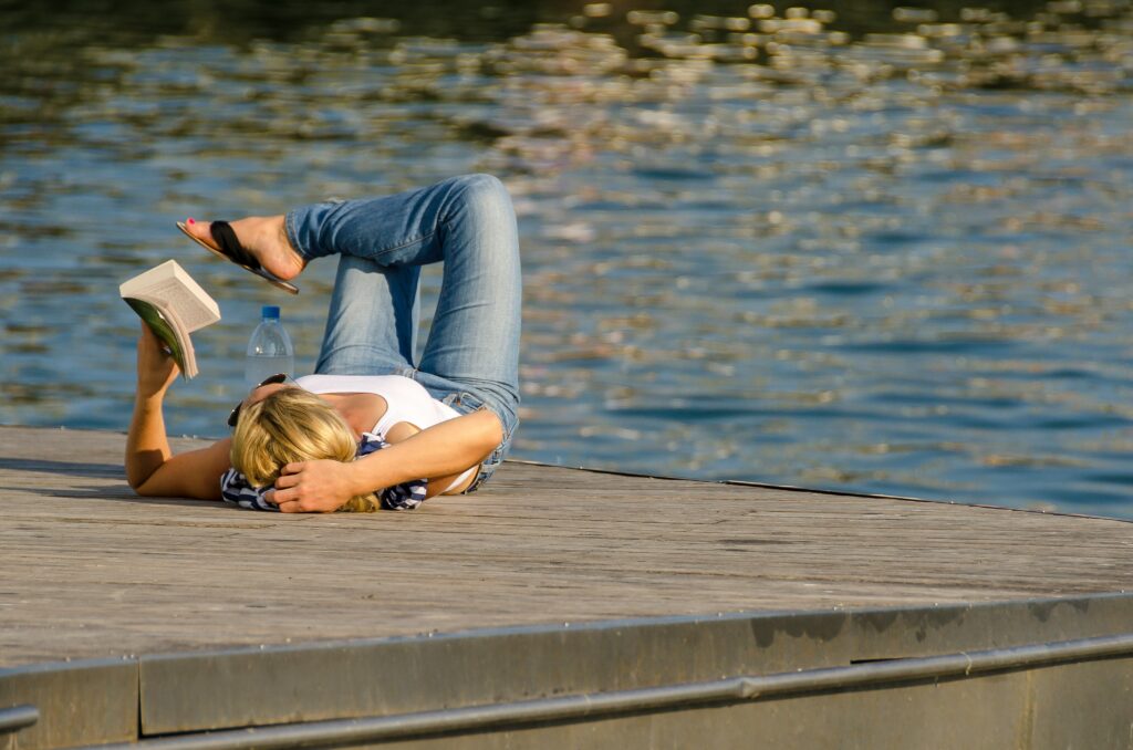 a woman laying down and reading a book near a lake 