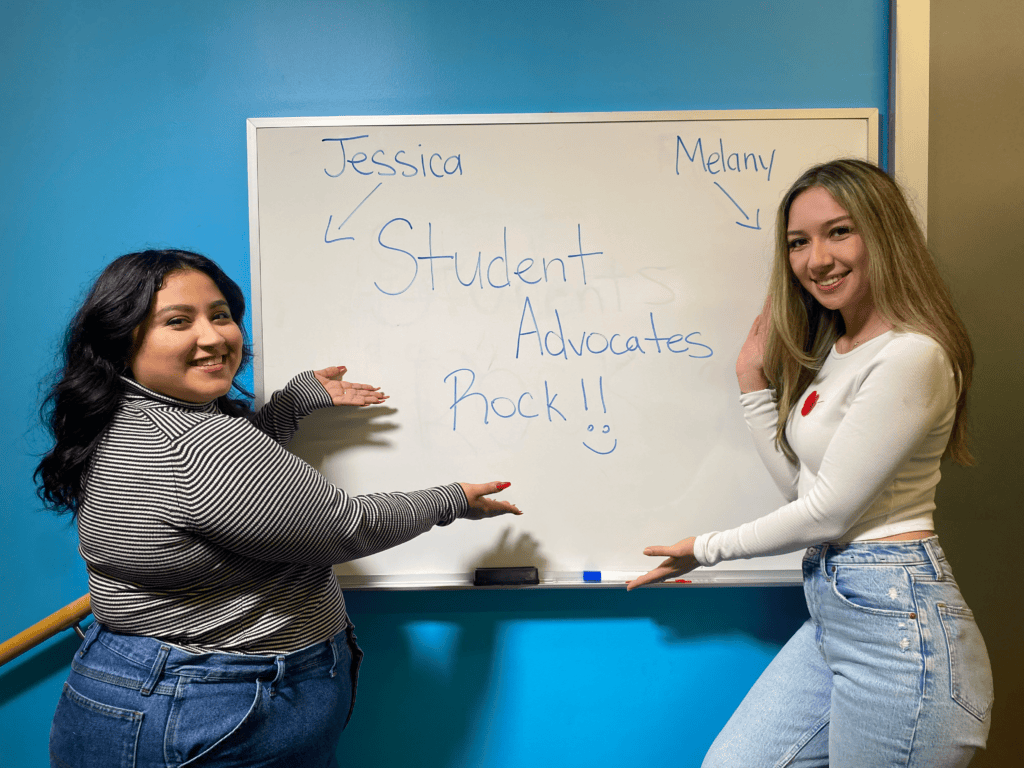 Melany and Jessica are standing next to a white board and smiling. (honest)