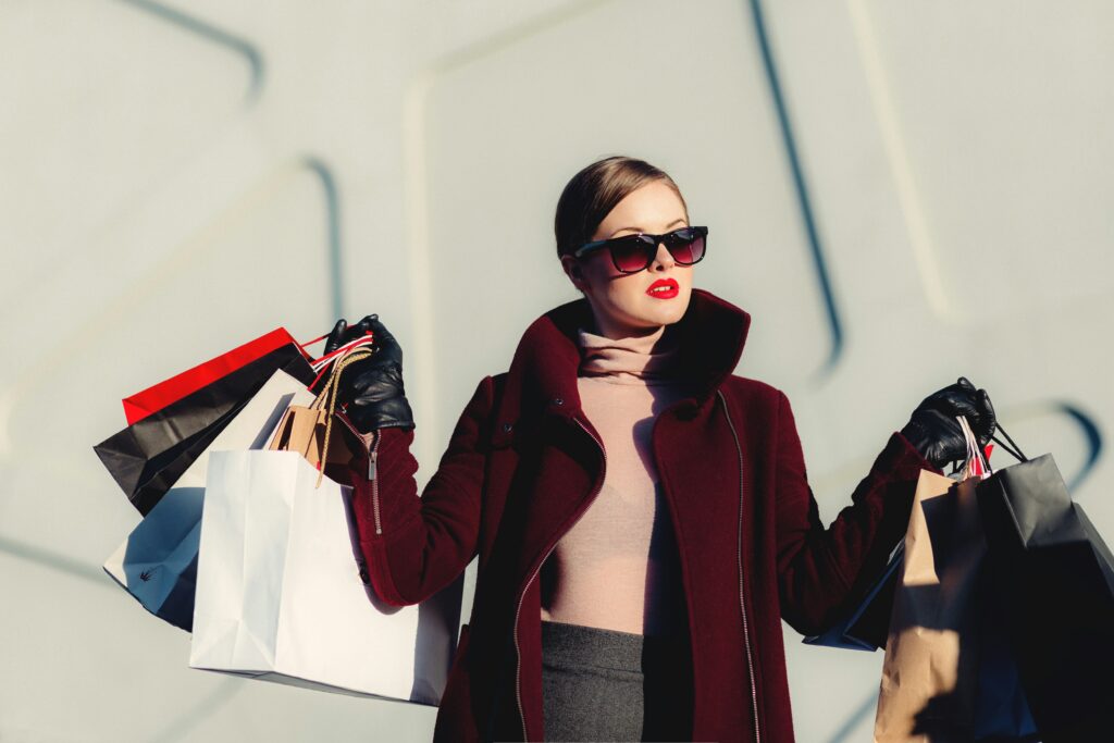 A person holding 6 shopping bags up in both her hands with red tinted sunglasses and a long maroon coat.
