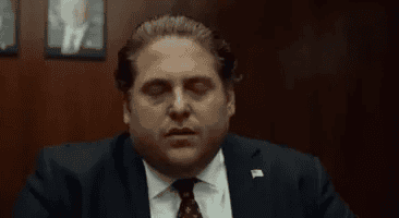 A man in a suit with slicked-back hair is sitting with his eyes closed, appearing stressed or frustrated, in a wood-paneled room with framed photos on the wall behind him.