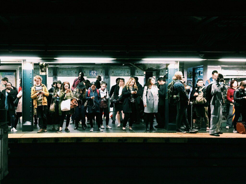 A group of people stand on a subway platform, some checking phones or talking, waiting for a train. Fluorescent lights illuminate the scene. The mood appears busy and urban.