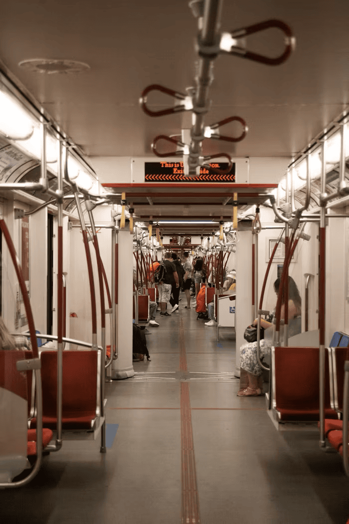 Passengers sit and stand inside a subway train with red seats and overhead handrails, while an electronic sign displays route information at the far end of the car.
