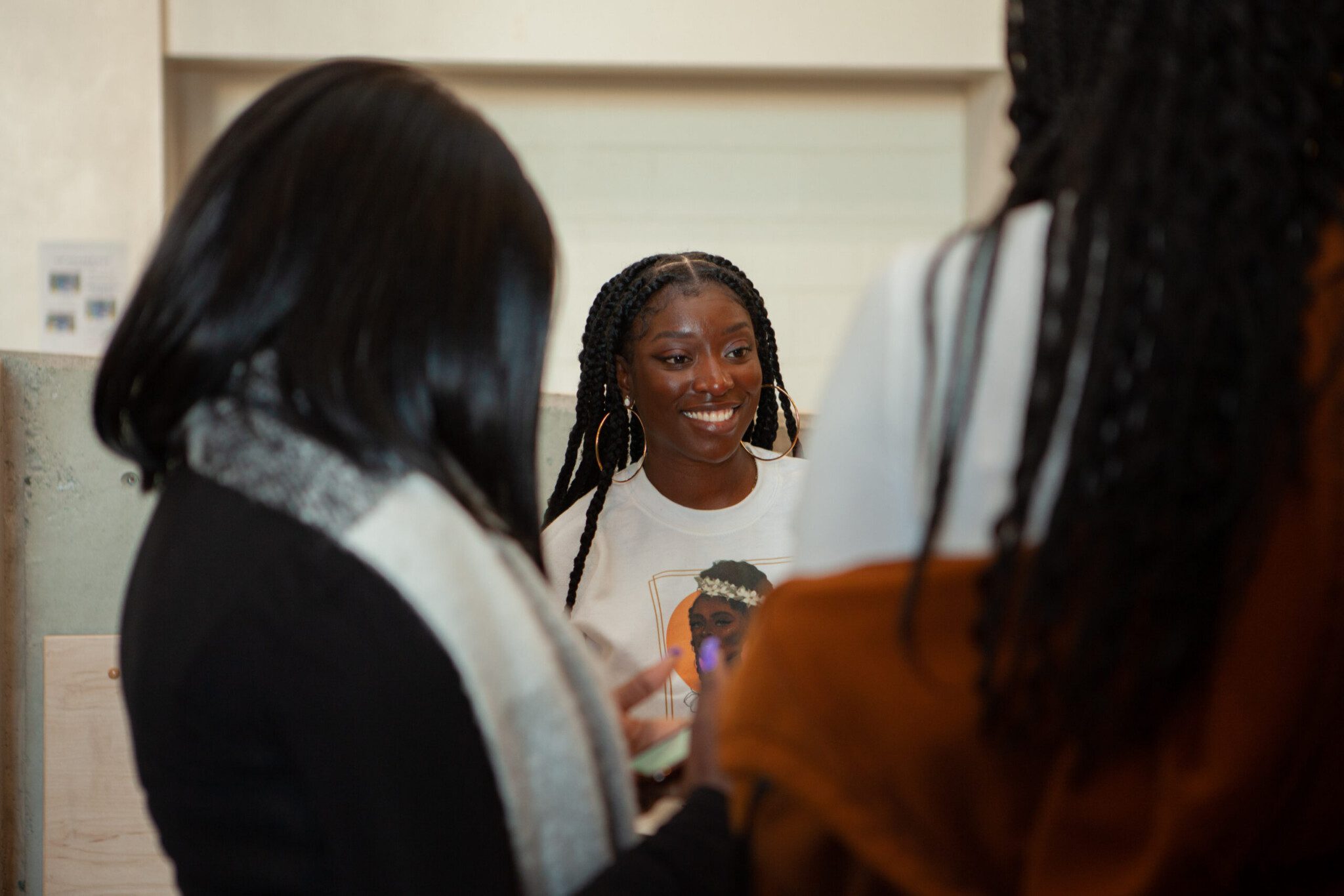Smiling woman with braided hair in a light shirt, surrounded by people indoors.