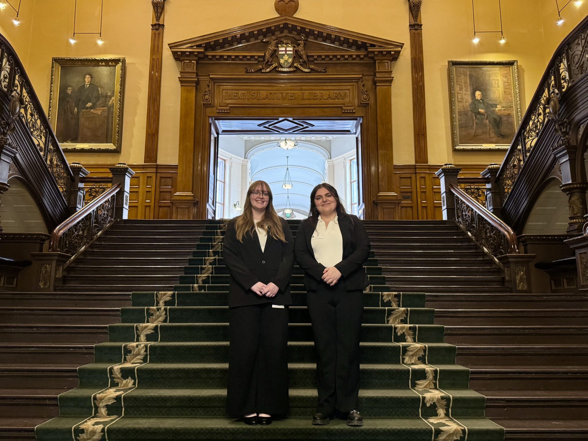 Two women in black suits pose on a grand wooden staircase inside a historic legislative building, with framed portraits on the walls and ornate iron railings on either side.