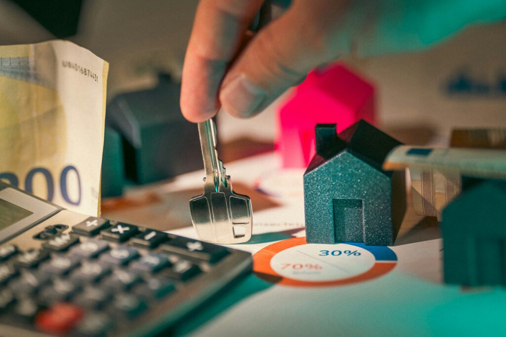 A person's hand holding a key upright on a table with one finger at its point with money and miniture houses around it. 