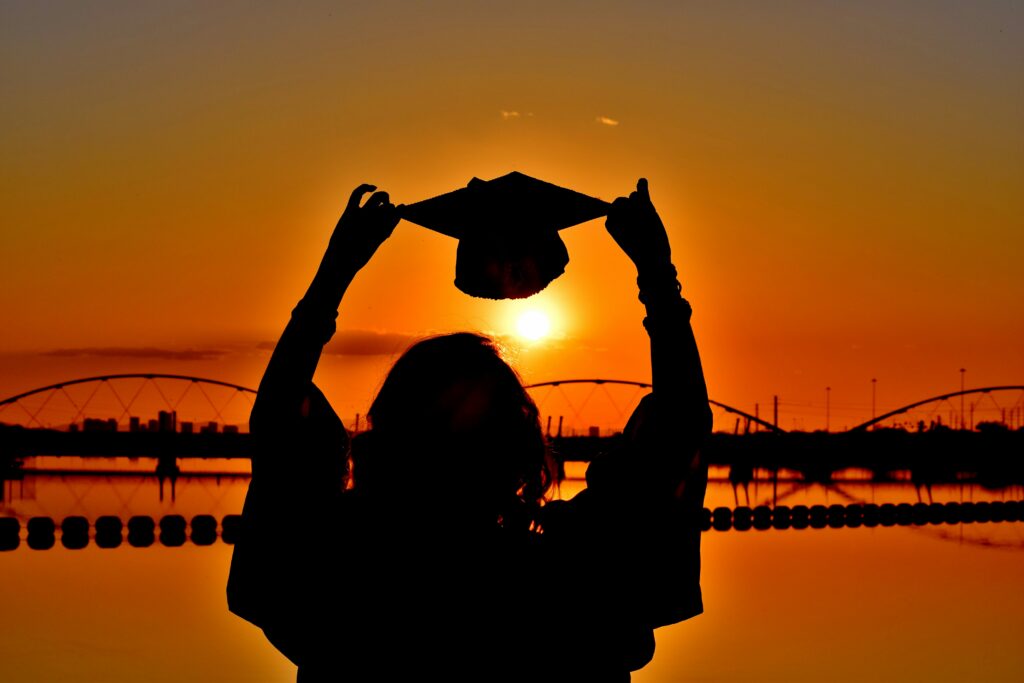 A person holding up a graduation cap, looking over a waterfront with the silhouette of a bridge in the distance.