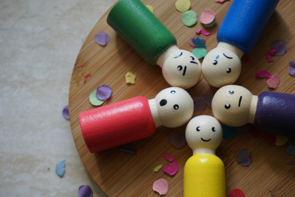 Five wooden figures with different colored bodies and various facial expressions lie in a circle on a wooden surface, surrounded by colorful confetti.