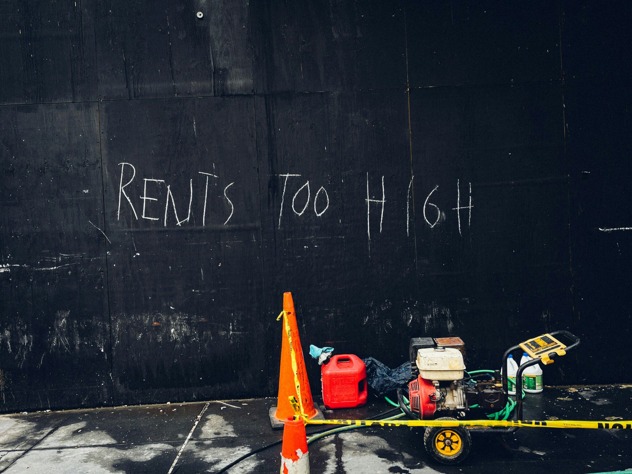 A black wooden construction wall featuring hand-painted white graffiti that reads "RENTS TOO HIGH," with an orange traffic cone, red gas can, and portable generator sitting on the sidewalk below.