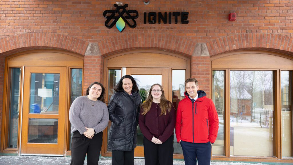 Four people stand smiling in front of a brick building with an "IGNITE" sign above the entrance.