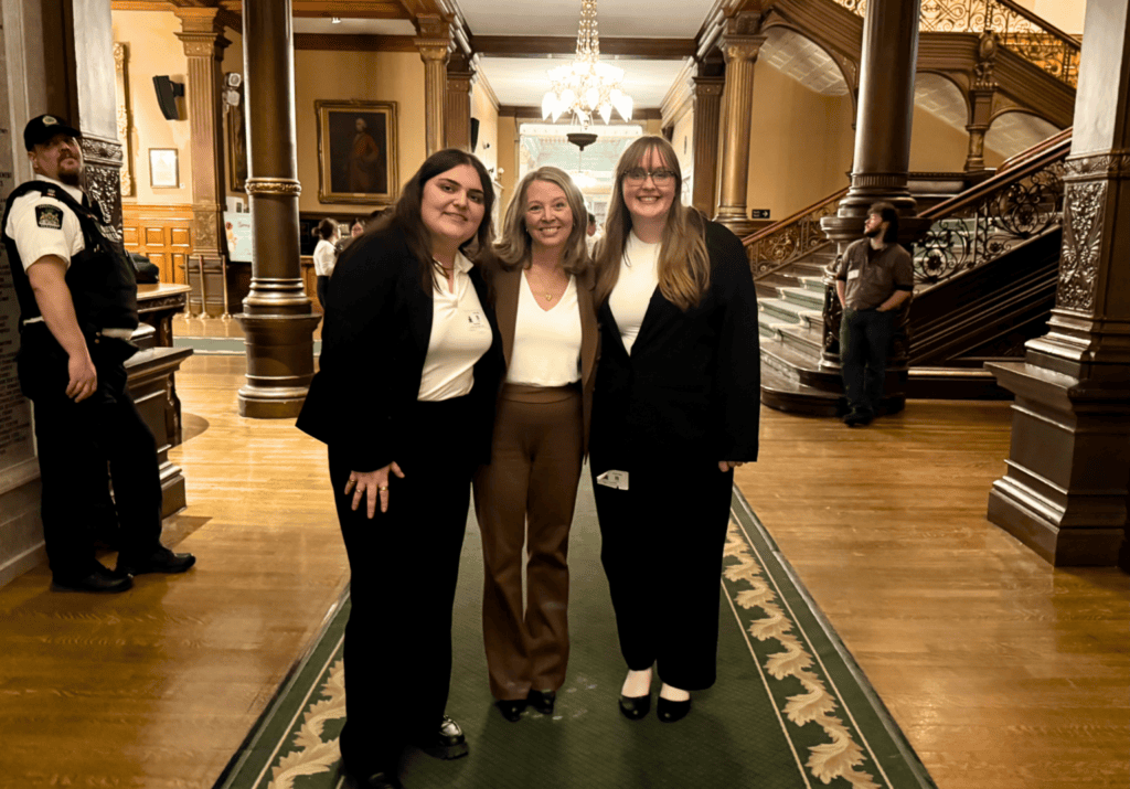 Three smiling women in business attire pose together in a grand historic lobby with wooden columns and a crystal chandelier. A security guard stands to the left.