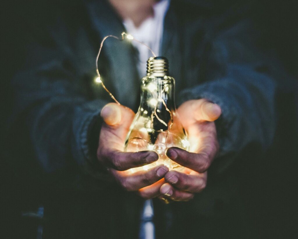 Two hands holding an upside-down illuminated bulb with illuminated wires running out from underneath it.