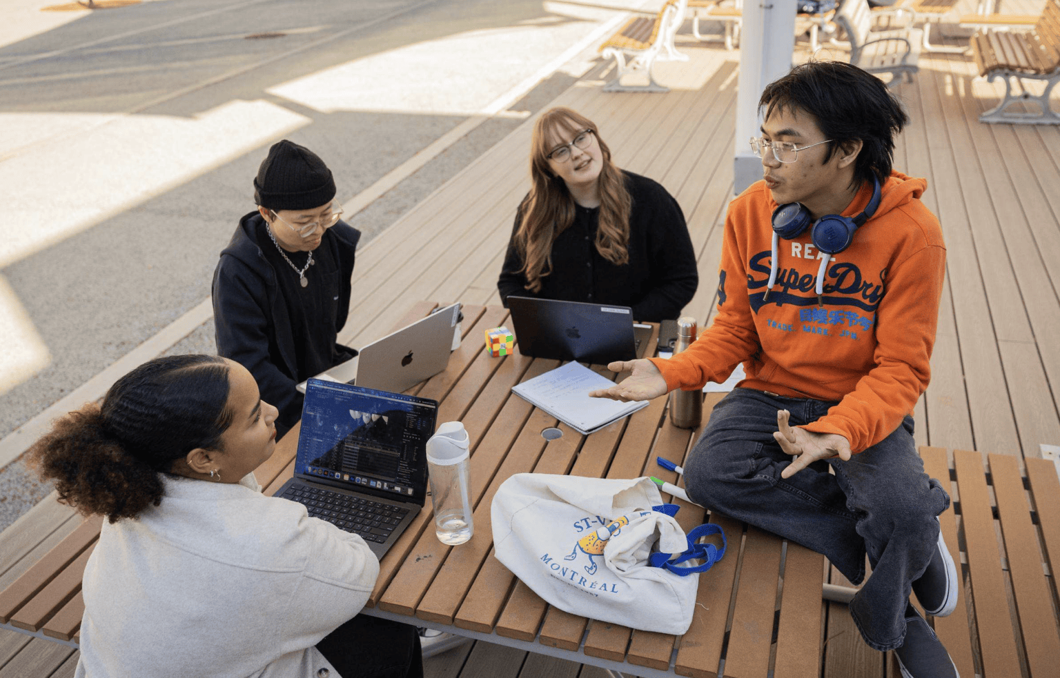 Group of diverse students seated around a wooden picnic table outdoors, collaborating with laptops and notes.