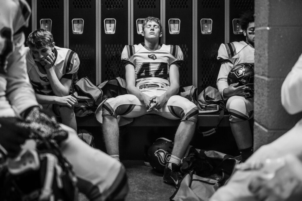 A football athlete sitting on a bench in a locker room with his teammates with his eyes closed leaning back against lockers and his hands entwined.