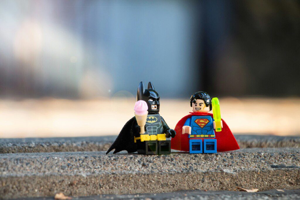 Two LEGO superheroes, Batman and Superman, stand on a rough outdoor surface with capes; Batman holds a pink ice cream cone.