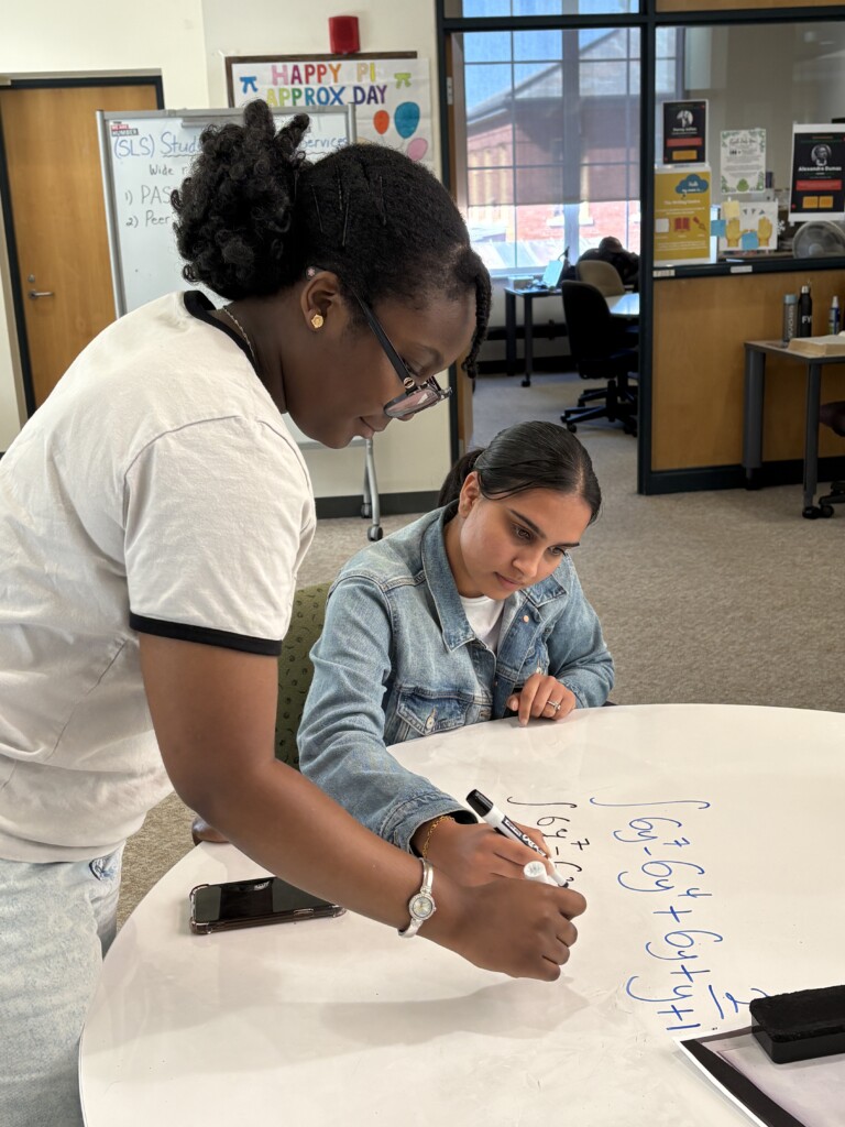 A tutor and a student writing out a math equation in marker on a white desk.