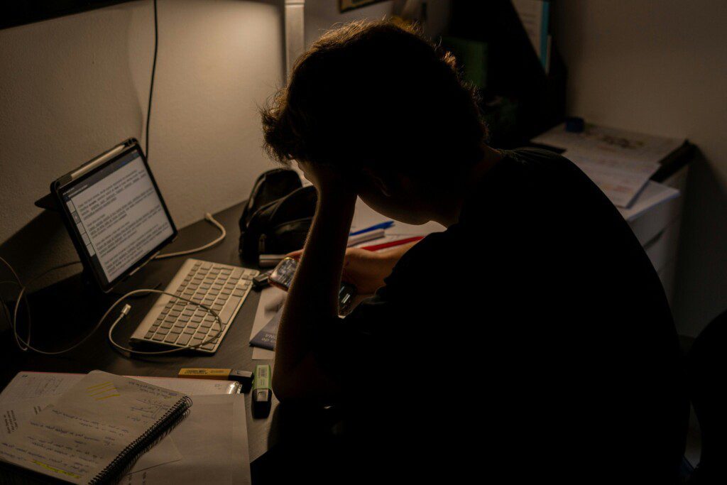 A person sitting in front of a computer and looking on his phone with papers and school supplies scattered on the desk around him