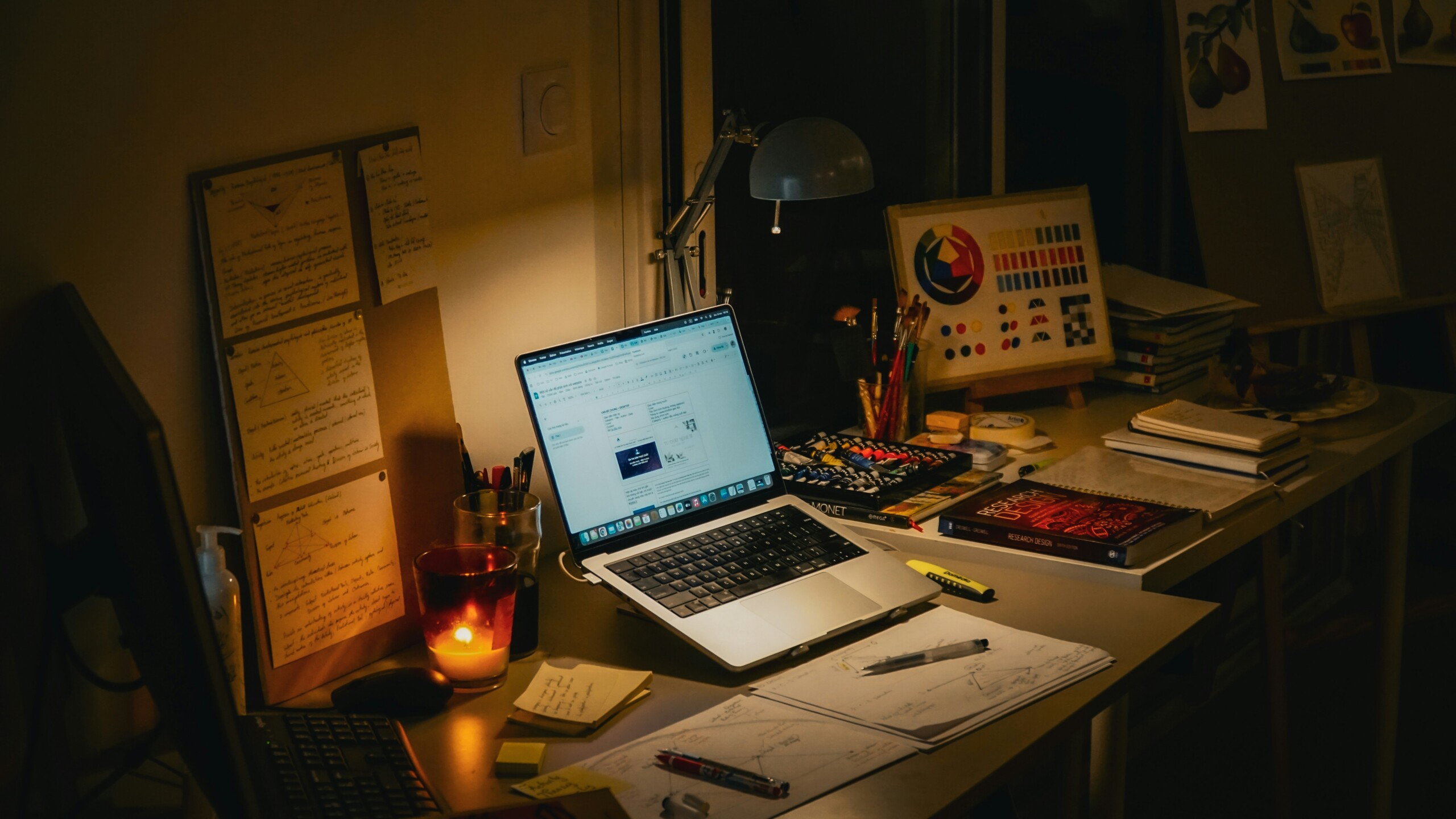 A desk in dim lighting, cluttered with papers and art supplies surrounding a laptop