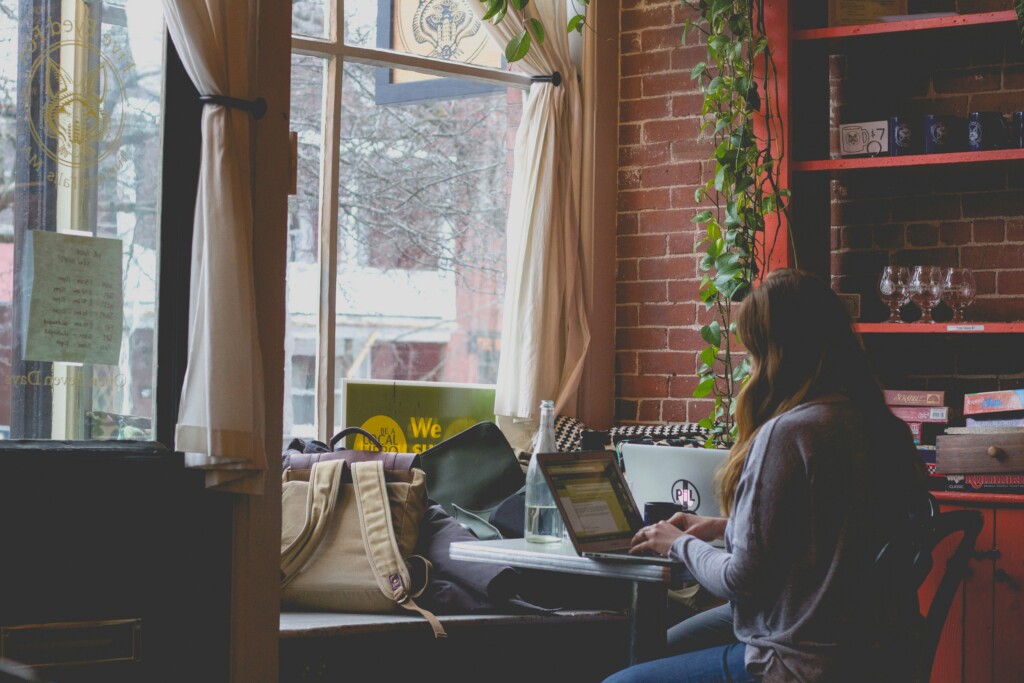 woman with long brown hair using a laptop in a cafe with brick walls and large windows.
