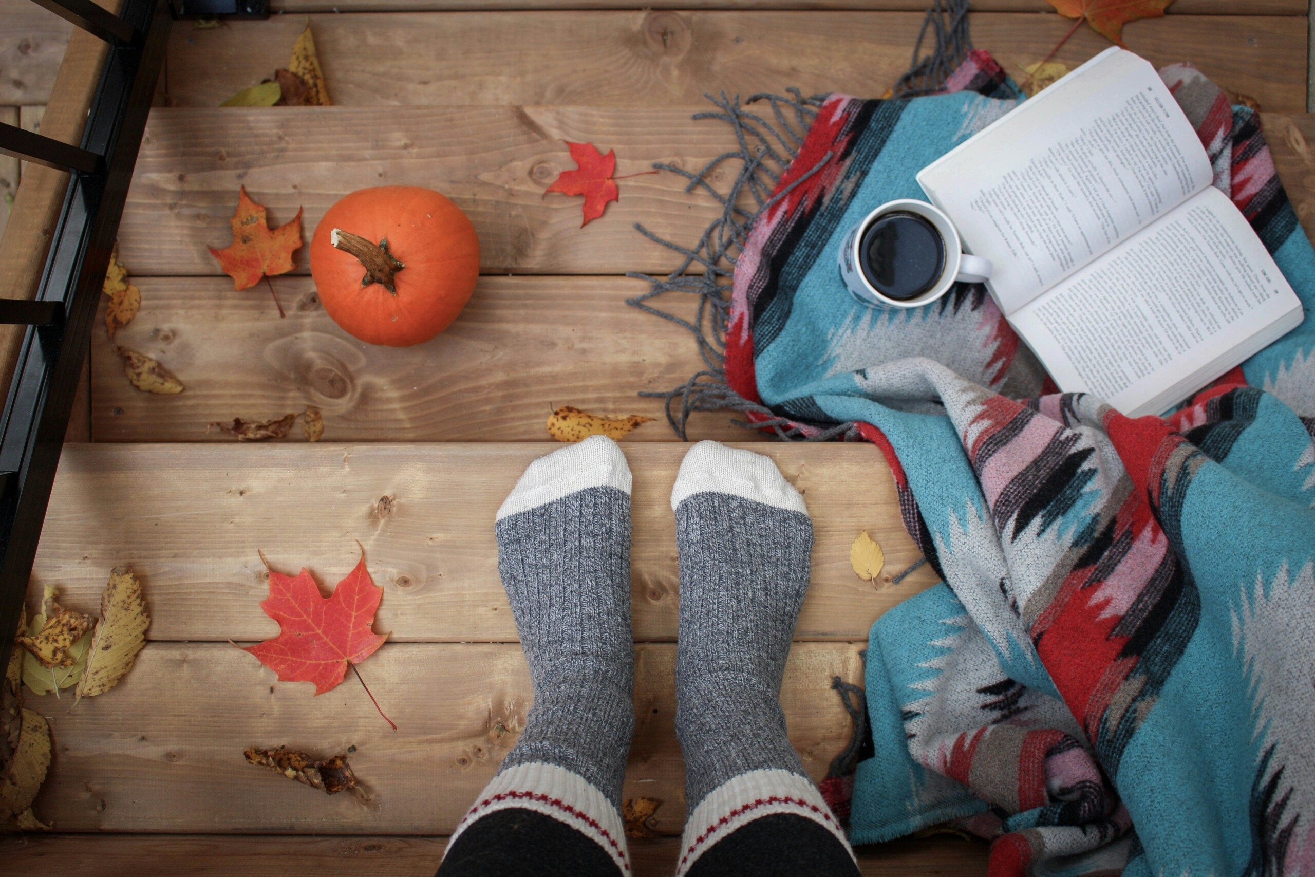 Two feet wearing grey socks standing on wooden stairs, beside fall leaves, a small pumpkin, a cup of coffee and an opened book on a blanket